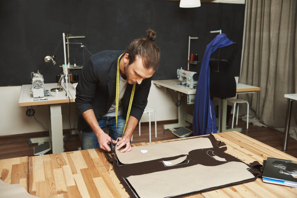 mature creative handsome dark haired caucasian male fashion designer in black suit cutting out clothes parts from fabric with scissors, spending evening in studio.